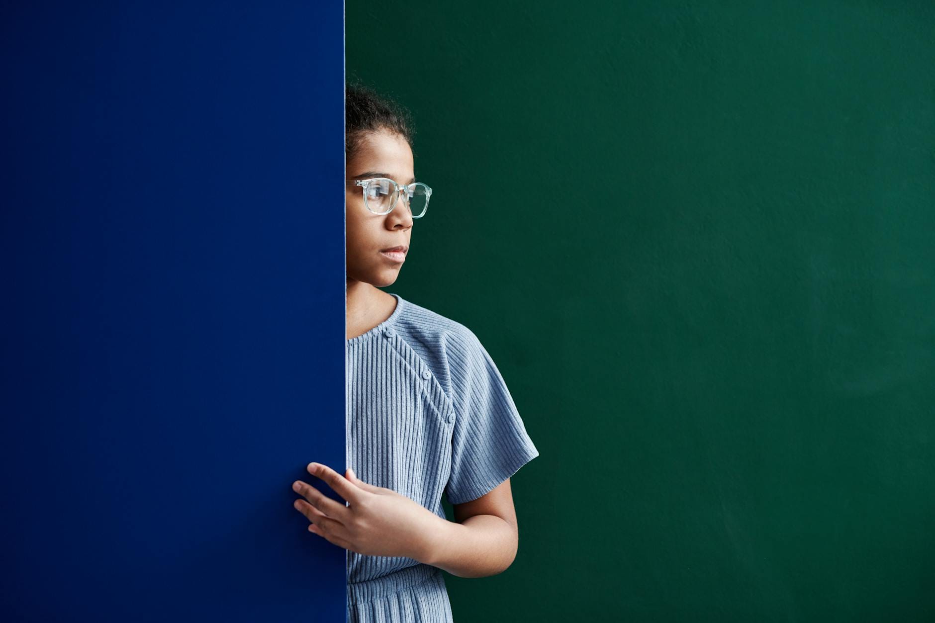 Zamyślona nastolatka w okularach stoi przy niebieskiej szkolnej ścianie