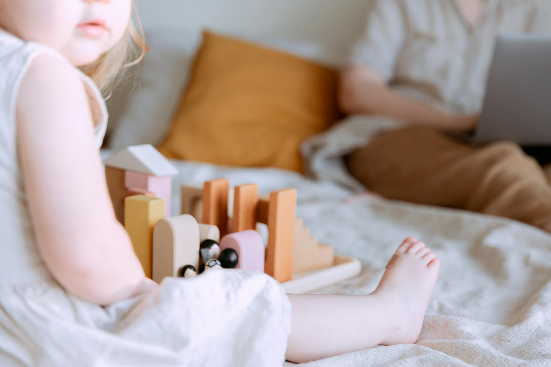 Mama pracuje zdalnie na łóżku, obok córka bawi się drewnianymi klockami